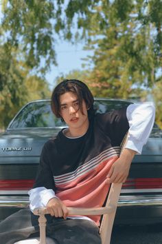 a young man sitting in a chair next to a parked car and looking at the camera