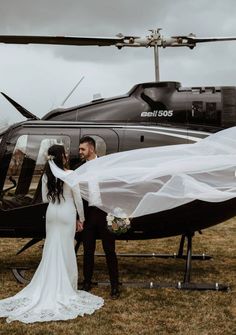 a bride and groom standing in front of a helicopter with their veil blowing in the wind