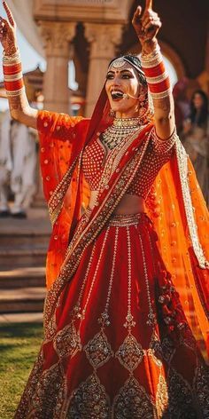 a woman in an orange and red bridal outfit with her hands up to the sky