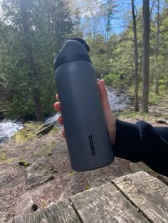 a person holding a water bottle in their hand near a stream and trees on the other side