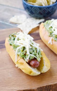 two hot dogs covered in guacamole and cheese on a wooden cutting board