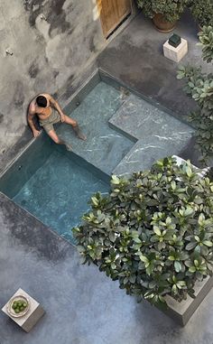 a man sitting on the edge of a swimming pool surrounded by greenery and potted plants