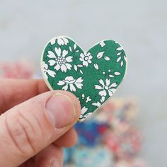 a hand holding a green and white heart shaped brooch with daisies on it