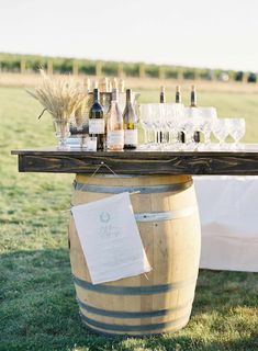 a table with wine glasses and bottles sitting on top of a barrel in the grass