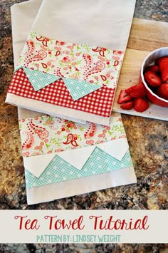 two tea towels with red strawberries in a bowl next to them on a cutting board