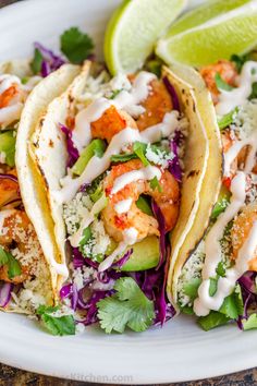 three shrimp tacos on a plate with lime wedges and cilantro sauce