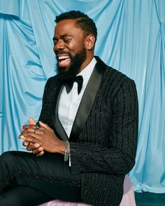 a man in a tuxedo laughing and sitting on a pink chair with blue curtains behind him