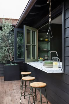 three stools sit on a wooden deck near an outdoor kitchen area with black walls and windows