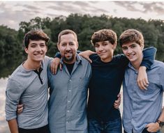 four young men standing next to each other in front of a body of water with trees in the background