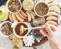 a person holding a wooden spoon over an assortment of food on a platter with apples, marshmallows and chocolate