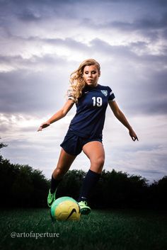 a young woman kicking a soccer ball on top of a lush green field in front of a cloudy sky