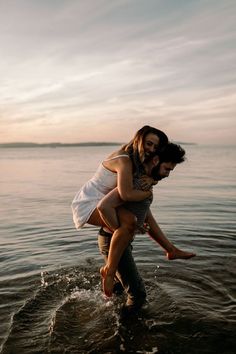 a man holding a woman in his arms as they walk through the water at sunset