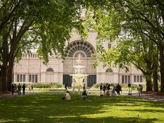 people are sitting on the grass in front of a building with a fountain and trees