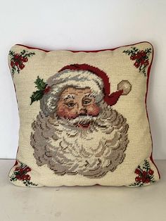 a cross stitch santa claus pillow on a white surface with red trimmings and holly berries