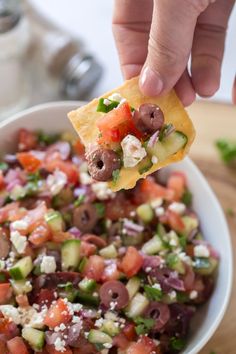 a hand holding a tortilla chip over a bowl filled with vegetables and cheese