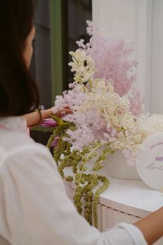 a woman arranging flowers in a vase on top of a white table next to a window