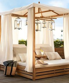 a canopy bed with white curtains and pillows on top of a wooden deck next to plants