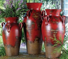 four red vases with plants in them on a table outside near some bushes and flowers