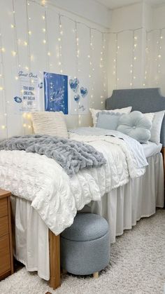 a bedroom with white bedding and fairy lights on the wall above it, along with a gray footstool