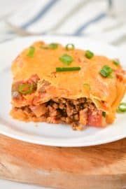 a white plate topped with lasagna covered in cheese and green onions on top of a wooden cutting board