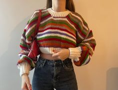 a woman wearing a multicolored sweater and jeans is standing in front of a white wall