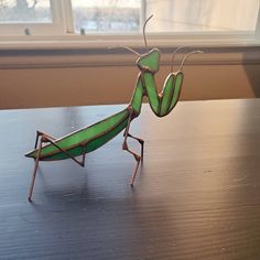 a green praying mantissa sitting on top of a wooden table next to a window