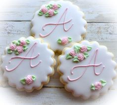 three decorated cookies with pink and green icing on a white wooden surface, one has the letter a