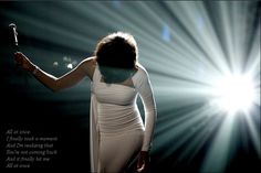 a woman in a white dress holding a microphone and standing on stage with bright lights behind her