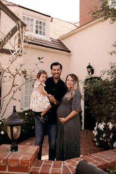 a man and woman standing in front of a house with their baby girl on her lap