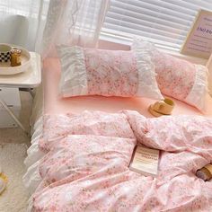 a bed covered in pink and white sheets with flowers on it, next to a window