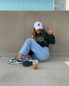 a woman sitting on the ground with her legs crossed and wearing a yankees hat, holding a starbucks cup