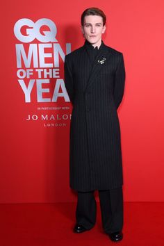 a man standing in front of a red wall wearing a long black coat and pants