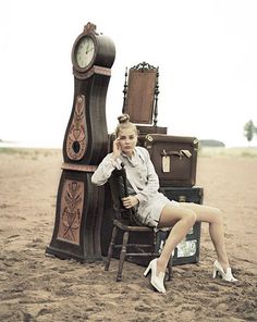 a woman sitting in a chair next to an old clock and suitcases on the beach