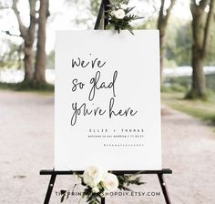 a welcome sign with flowers and greenery sits on an easel in front of trees