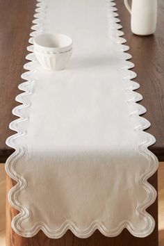 a white table runner with scalloped edges on a wooden table next to a vase