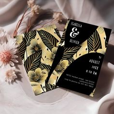 two black and gold wedding cards with floral designs on them next to a white flower