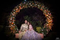 a man and woman sitting on top of a bench in front of a floral arch