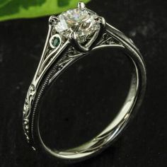 a white gold engagement ring with two green emeralds on the sides and an old style filigree setting