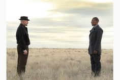 two men standing in the middle of a dry grass field with one man wearing a suit and tie