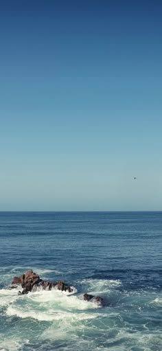 an airplane flying over the ocean with waves crashing on it's shore and rocks in the foreground