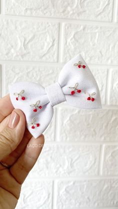 a hand holding a small white bow with red flowers on the front and side, in front of a white brick wall