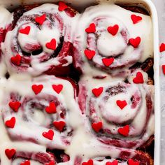 a white dish filled with cinnamon rolls covered in icing and red heart shaped candies