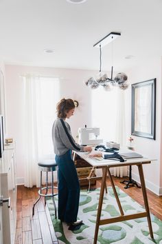 a woman standing at a sewing machine on a table in front of a large window