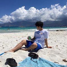 a man sitting on top of a sandy beach next to the ocean holding a drink