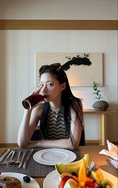 a woman sitting at a table drinking from a bottle with fruit on the plate next to her