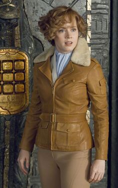 a woman in brown leather jacket standing next to a wall with gold decorations on it