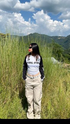 a woman standing on top of a lush green field next to tall grass and mountains