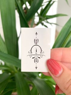 a person holding up a small card with a design on it in front of a potted plant