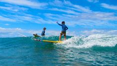 two people riding surfboards on top of a wave in the ocean with blue sky and clouds