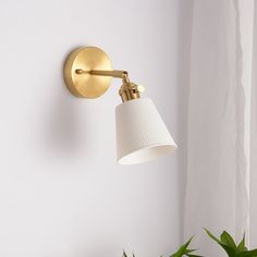 a wall light with a white shade hanging from it's side next to a potted plant